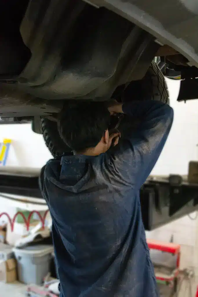 A vehicle repair shop employee standing underneath a lifted car making repairs to a tire